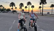 Two people are smiling and posing on their bicycles during sunset by a palm-lined waterfront.