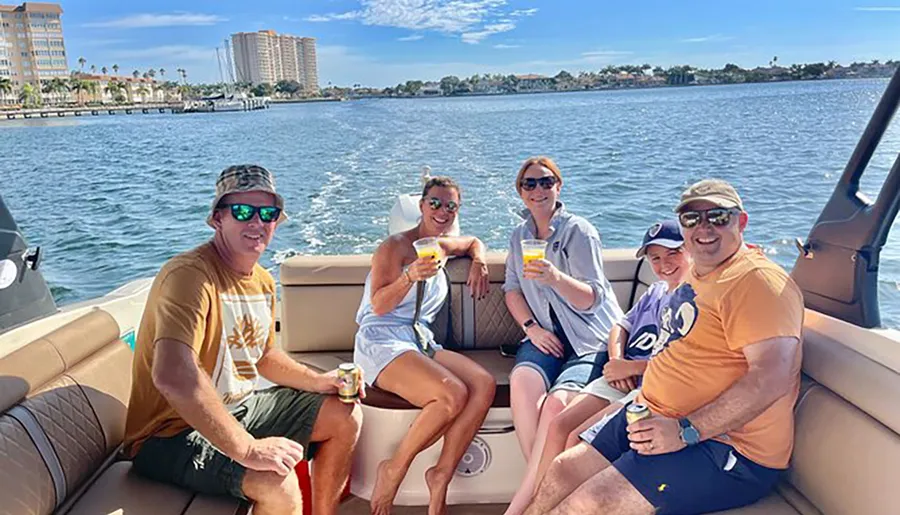 A group of adults and a child are enjoying a sunny day on a boat, with some holding drinks, against a backdrop of calm water and coastal buildings.
