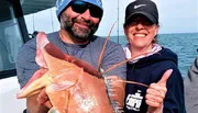 A man and a woman are smiling on a boat, proudly displaying a large, reddish fish they have caught.