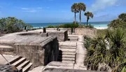 The image depicts an old concrete military fortification overlooking a tropical beach with palm trees and a clear blue sea.