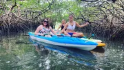 Three people are smiling and enjoying a kayaking excursion through a calm waterway surrounded by dense mangrove trees.