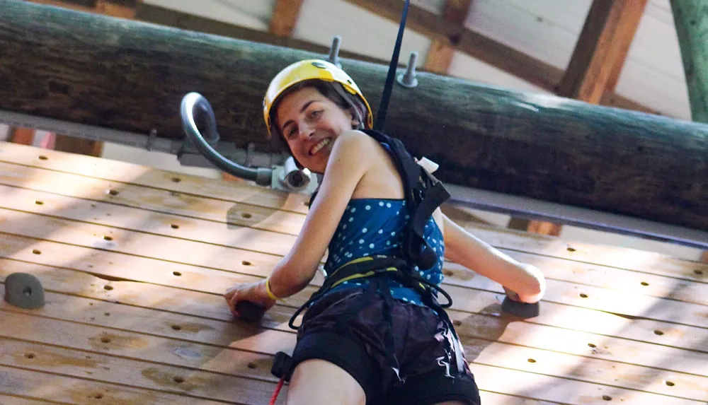 A person wearing a climbing harness and safety helmet is smiling while ascending an indoor climbing wall