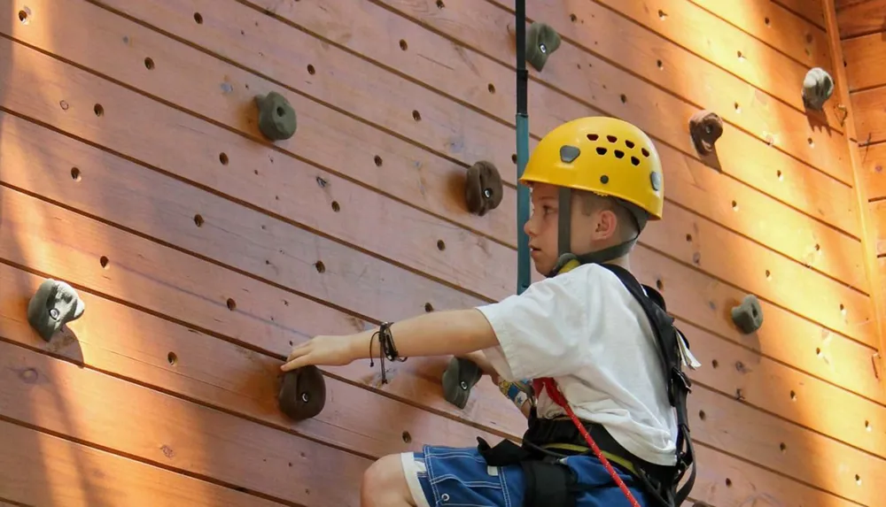 A young person wearing a helmet and harness is climbing an indoor rock wall