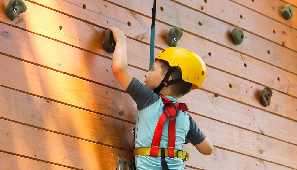 A child wearing a safety harness and helmet is climbing an indoor rock wall