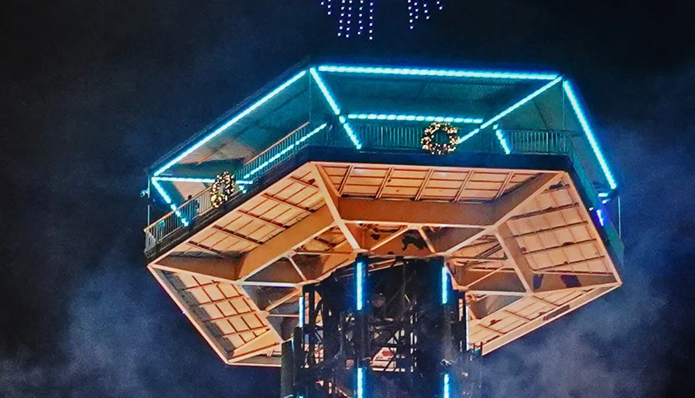 This image features a close-up of the illuminated top section of a tower at night with blue lights outlining its structure and adding a futuristic or festive touch