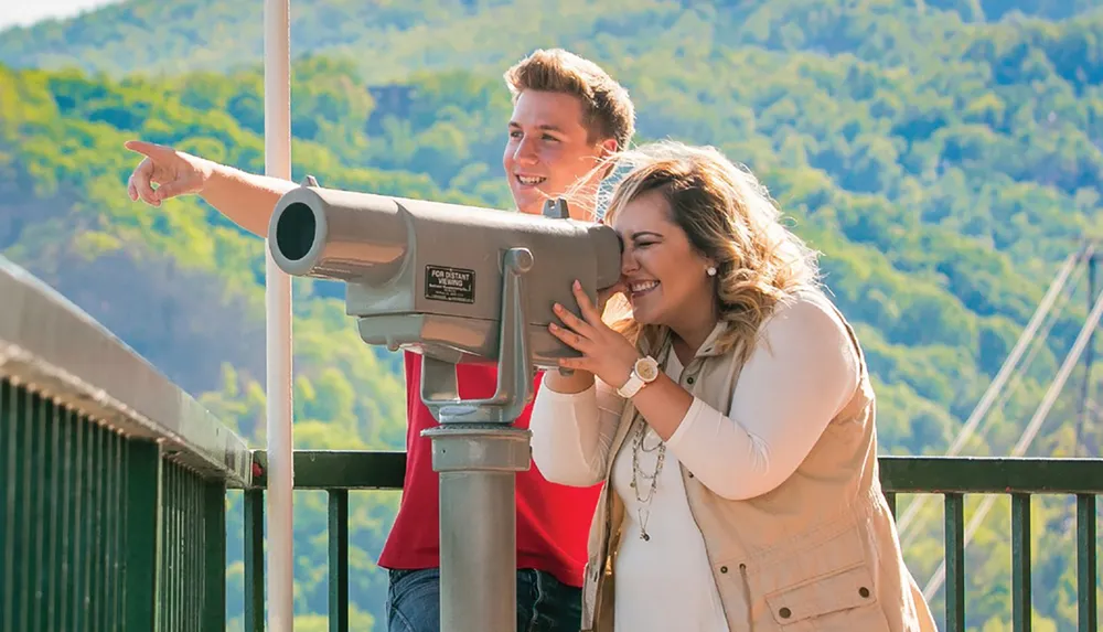 A joyful couple engages with a coin-operated telescope at a scenic overlook with the man pointing out a sight while the woman looks on laughing