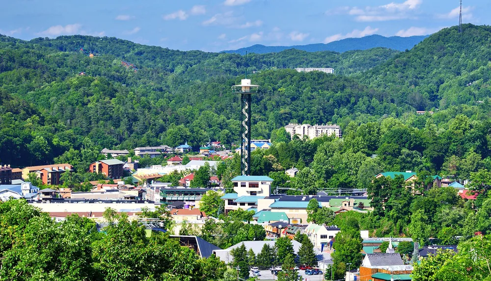 The image depicts a vibrant town nestled among lush green hills with a prominent observation tower standing tall amidst the buildings under a blue sky scattered with clouds