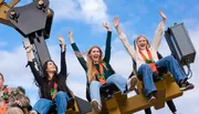 Thrilled amusement park guests are raising their hands high while enjoying the exciting motion of a ride against a blue sky.