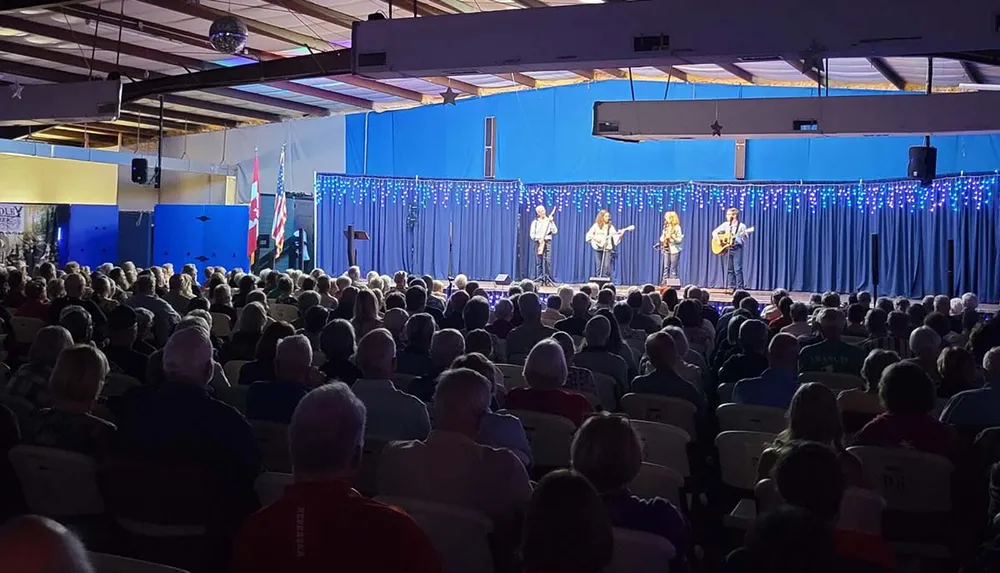 A band performs on stage before an attentive audience in a community hall adorned with blue curtains and festive lights