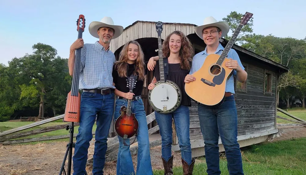 Four individuals possibly a family stand in front of a rustic building each holding a musical instrument suggesting they are a country or folk music band