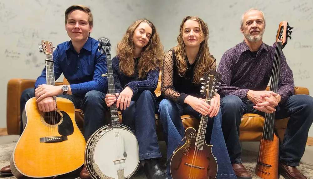 Four musicians are smiling and seated with stringed instruments including a guitar banjo mandolin and a stand-up bass against a backdrop of a wall covered in signatures