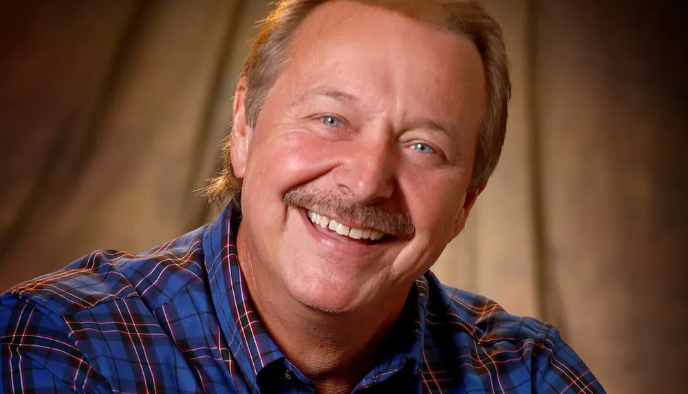 A smiling man with a mustache wearing a plaid shirt is against a blurred brown background