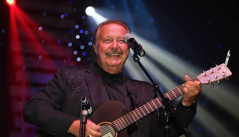 A smiling musician is playing the guitar on stage with colorful lights in the background