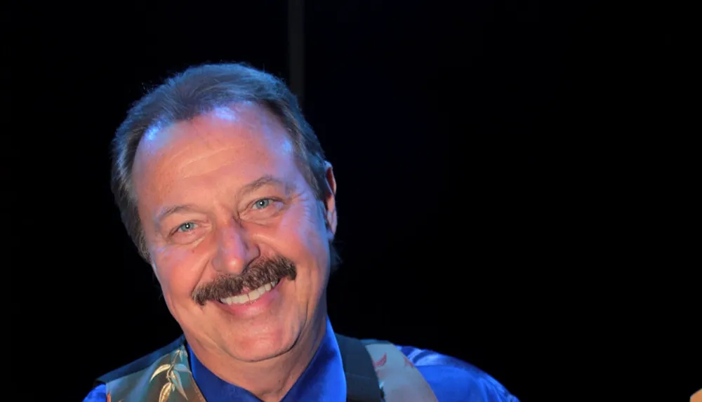 A man with a mustache is smiling at the camera against a dark background