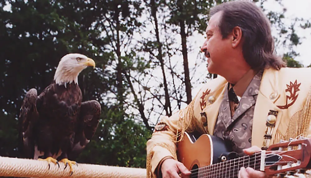 A man in a decorated jacket plays guitar next to a perched bald eagle in a natural outdoor setting