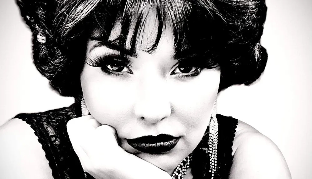 The image is a high-contrast black-and-white close-up portrait of a woman with vintage-styled makeup and hair gazing into the camera as she rests her chin on her hand