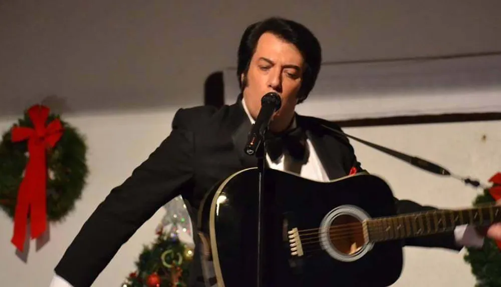 A person dressed in a black suit is performing onstage with a guitar and a microphone with Christmas decorations in the background