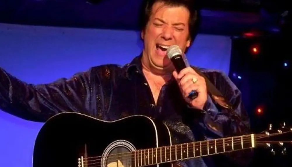 A person is passionately singing into a microphone with a guitar in the foreground and colorful lights in the background