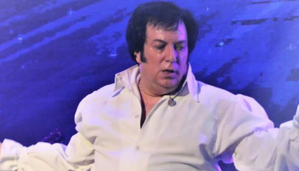 A person dressed in white is sitting with a focused expression possibly during a performance or event with a blue-lit background