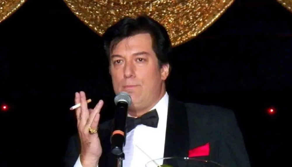 A man in a black tuxedo and bow tie is holding a cigarette and a microphone with a glittery background