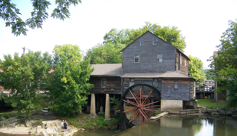 The image shows a rustic weathered wooden mill with a large red water wheel reflected in the tranquil water of a pond surrounded by lush greenery and two individuals standing by the waters edge