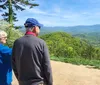 Two people are standing on a trail enjoying a panoramic view of rolling green mountains under a clear blue sky