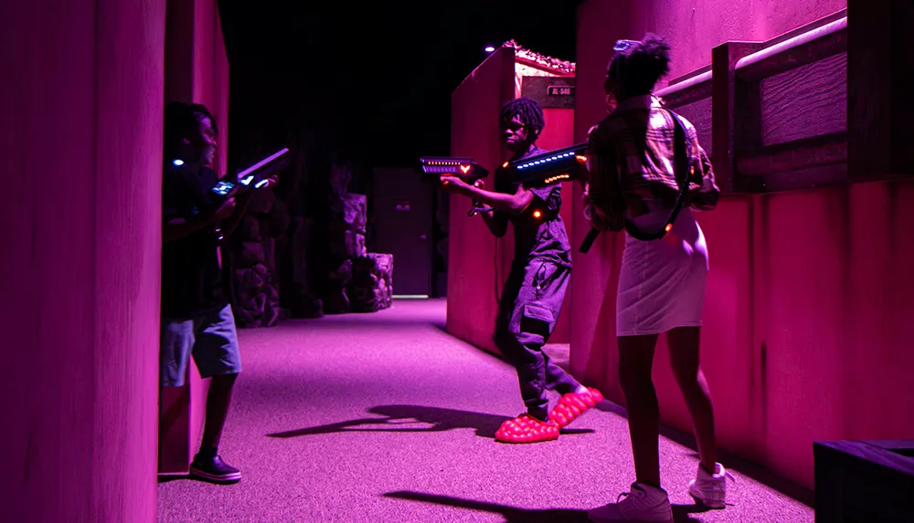 Three individuals are engaged in a laser tag game moving stealthily through a dimly lit arena with illuminated laser guns at the ready