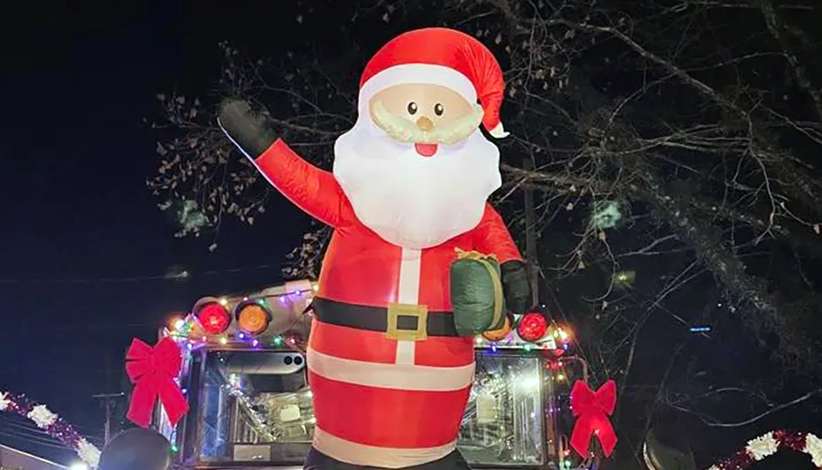 A large inflatable Santa Claus is positioned atop a festively decorated vehicle with Christmas lights and red bows at nighttime.