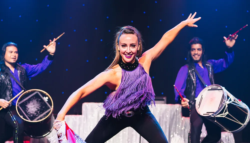 A dynamic performance with a woman dancing energetically in the foreground flanked by two drummers playing enthusiastically