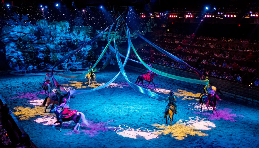 This image shows an indoor equestrian performance with riders in colorful costumes entertaining an audience in an arena with ambient lighting and dramatic decorations.