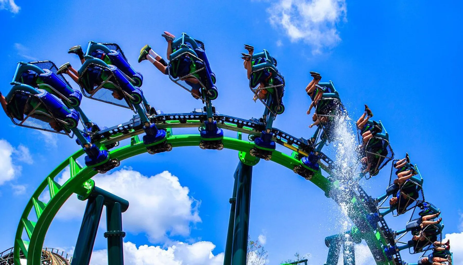 People are riding a looping roller coaster with green and blue tracks on a sunny day.