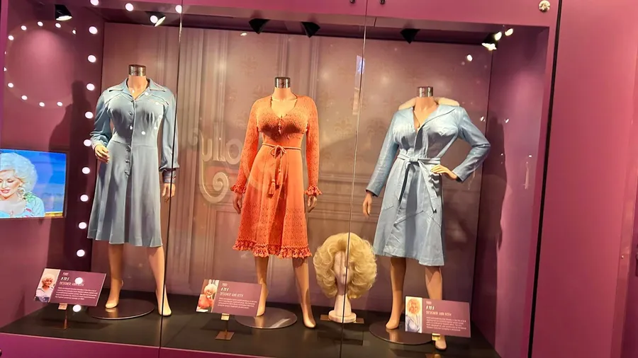 The image shows a display of three mannequins wearing vintage dresses with a blonde wig beneath one of the dresses, set against a purple background with a small screen and informational plaques.