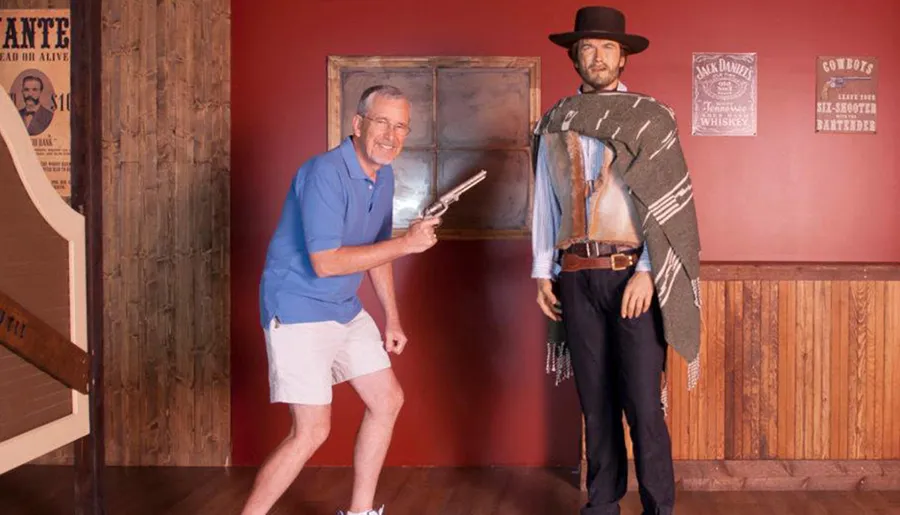 A person is playfully posing with a prop gun alongside a cowboy mannequin in a Wild West-themed setting.