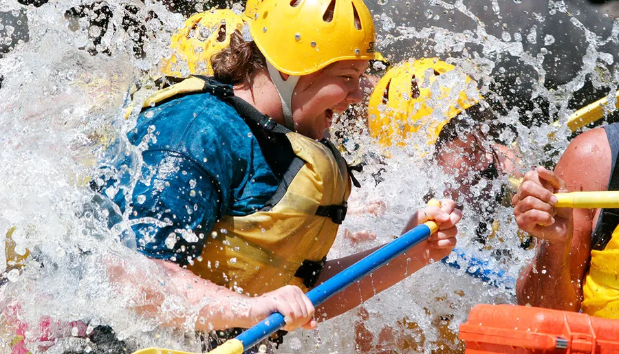 Two people in safety helmets and life vests are vigorously paddling through splashing water, likely engaged in a white-water rafting adventure.