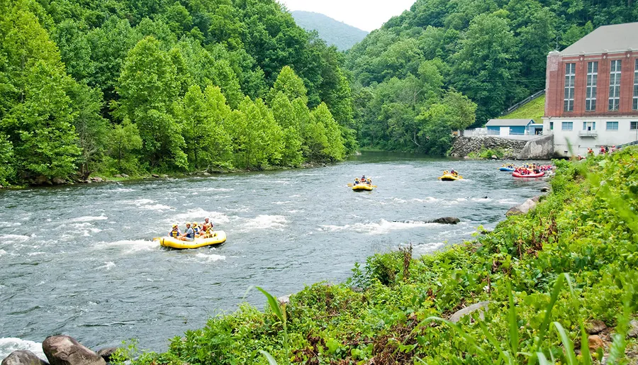 Groups of people are whitewater rafting down a scenic river surrounded by lush greenery and a building at the river's edge.