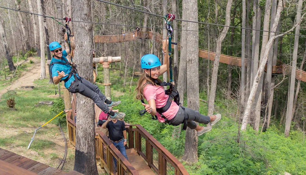 Three people are enjoying a zip-line adventure through a forested area with two of them actively gliding down the line while another waits on a platform