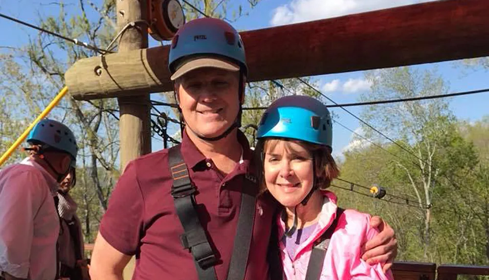 Two people wearing safety helmets and harnesses are smiling for a photo likely at an outdoor adventure or zipline park