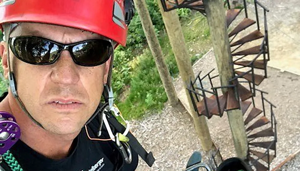 A person in a climbing helmet and harness takes a selfie at a high ropes course