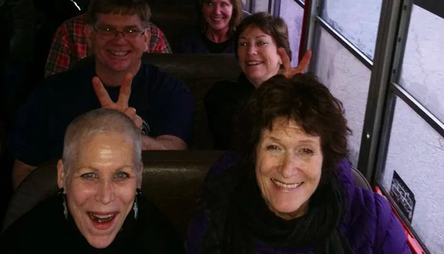 A group of cheerful people is posing for a photo on a bus, with one person making a playful hand gesture behind another's head.