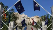 The image shows a view of the Mount Rushmore National Memorial with the carved faces of four American presidents, framed by an array of state flags.