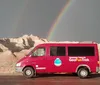 A red tour van is parked in front of a dramatic landscape with eroded rock formations under a dark sky with a double rainbow