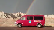 A red tour van is parked in front of a dramatic landscape with eroded rock formations under a dark sky with a double rainbow.