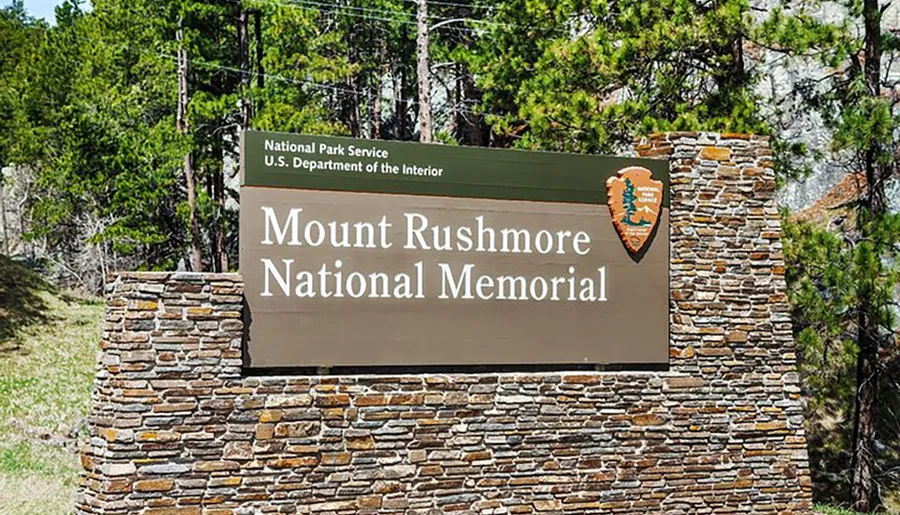The image shows the entrance sign for Mount Rushmore National Memorial, with the National Park Service emblem, indicating it is a site managed by the U.S. Department of the Interior.