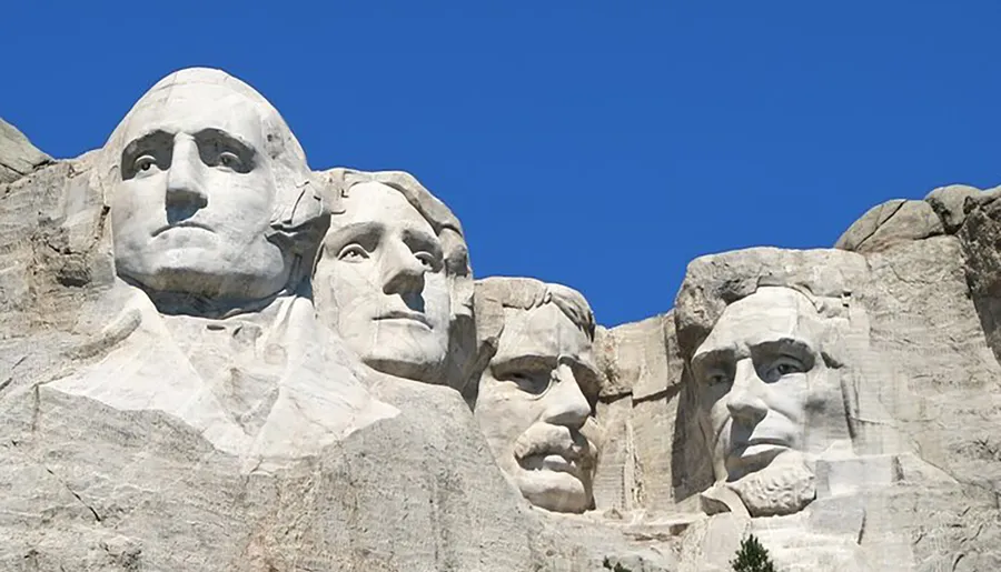 The image shows the iconic Mount Rushmore National Memorial, featuring the carved faces of four former U.S. presidents against a clear blue sky.