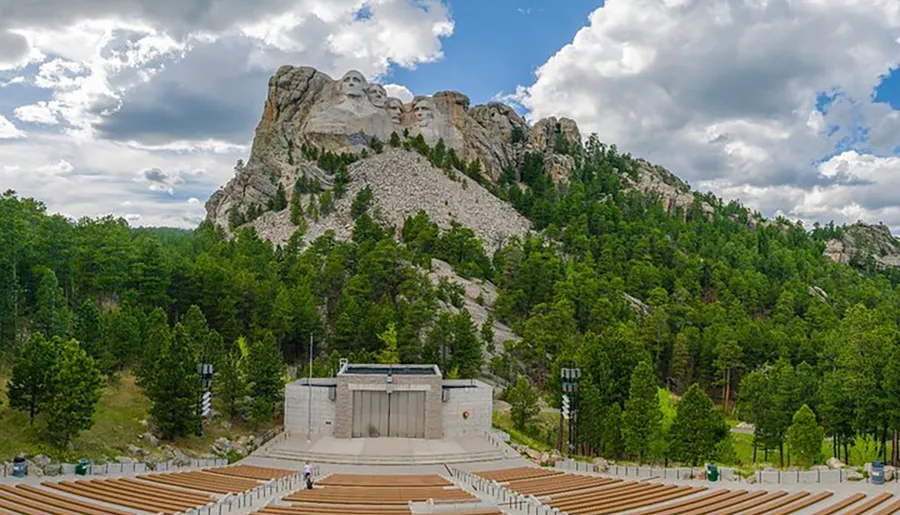 The image captures Mount Rushmore National Memorial, showing the colossal carvings of four former U.S. presidents' heads on a mountainside, with an amphitheater in the foreground set against a background of lush greenery and a partly cloudy sky.