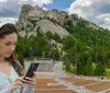 A young woman wearing headphones is looking at her smartphone in an amphitheater with the Mount Rushmore National Memorial visible in the background