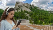 A young woman wearing headphones is looking at her smartphone in an amphitheater with the Mount Rushmore National Memorial visible in the background.