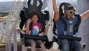 A child and an adult are experiencing different reactions while riding a thrilling amusement park attraction, with the child appearing apprehensive and the adult showing excitement.