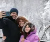 A family of three is taking a selfie in a snowy landscape all smiling and dressed warmly for winter weather