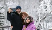 A family of three is taking a selfie in a snowy landscape, all smiling and dressed warmly for winter weather.